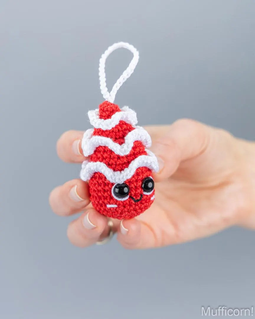 A hand holding a small crocheted chtistmas tree ornament, with a white loop at the top, against a soft gray background.
