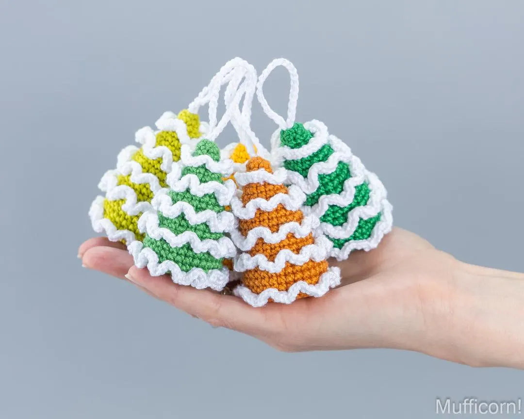 Three small crocheted Christmas tree ornaments in green, yellow, and orange with white ruffled trim are held in a person's hand against a plain gray background.