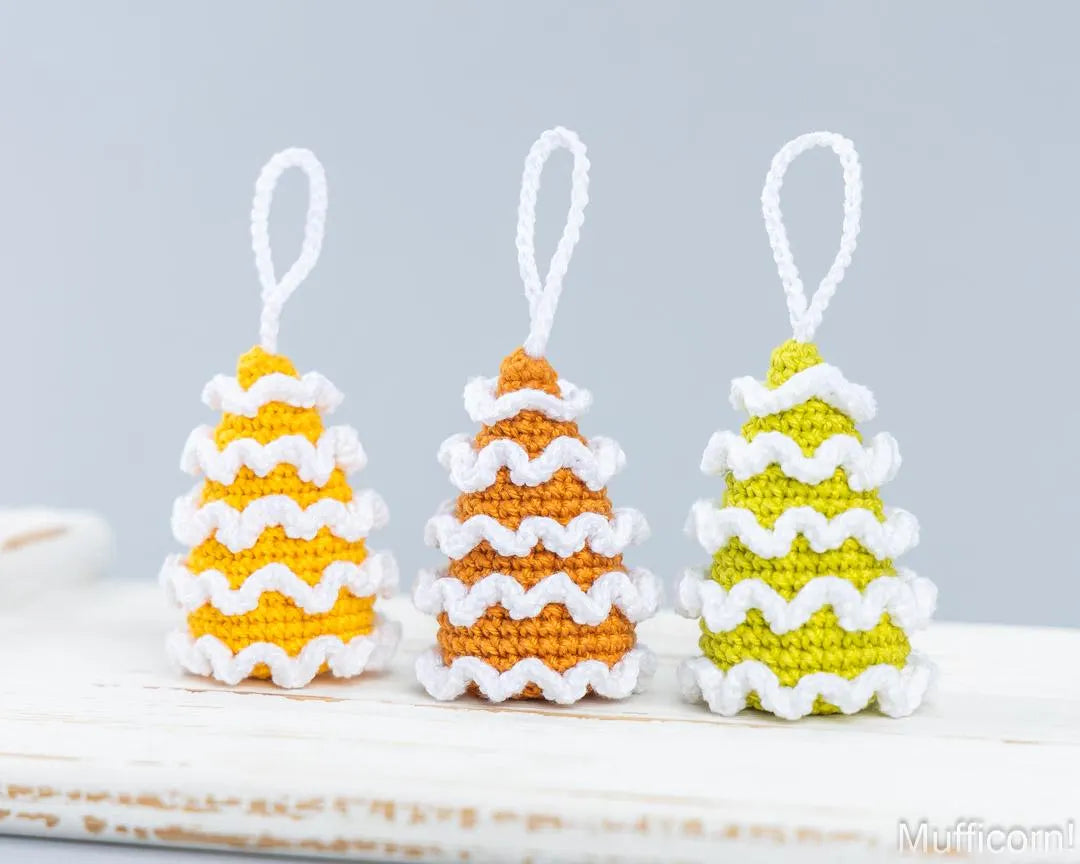 Three small crocheted Christmas tree ornaments with white hanging loops, shown on a white surface against a light gray background. The trees are yellow, orange, and green with white wavy trim.