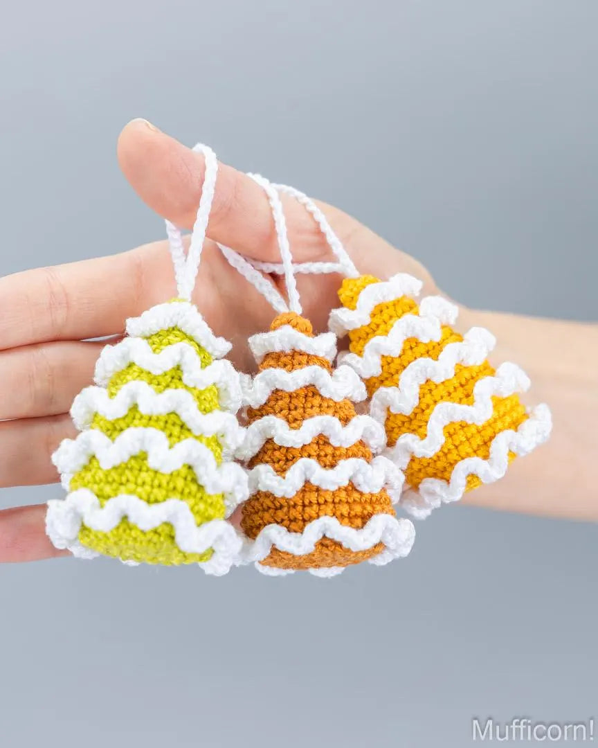 Three small crocheted bell-shaped christmas tree ornaments in yellow-green, orange, and yellow, each with a white zigzag trim and white hanging loops, held in a person's hand against a soft gray background.