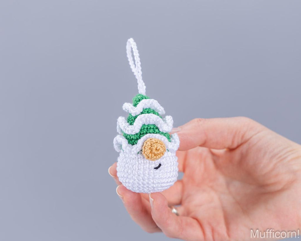A hand holding a small crocheted ornament shaped like a sleeping snowman with green and white textured hair, a gold button nose, and a white hanging loop, shown against a plain gray background.