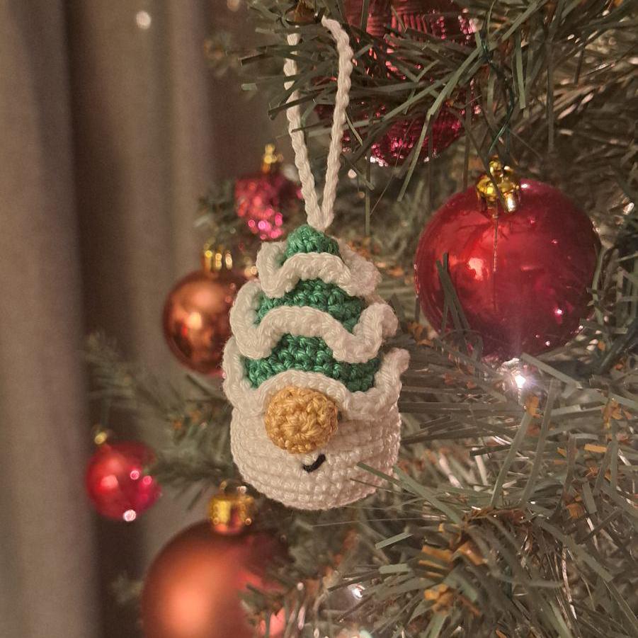 A crocheted Christmas ornament shaped like a snowman wearing a striped green and white winter hat, hanging on a decorated Christmas tree with red and gold ornaments.