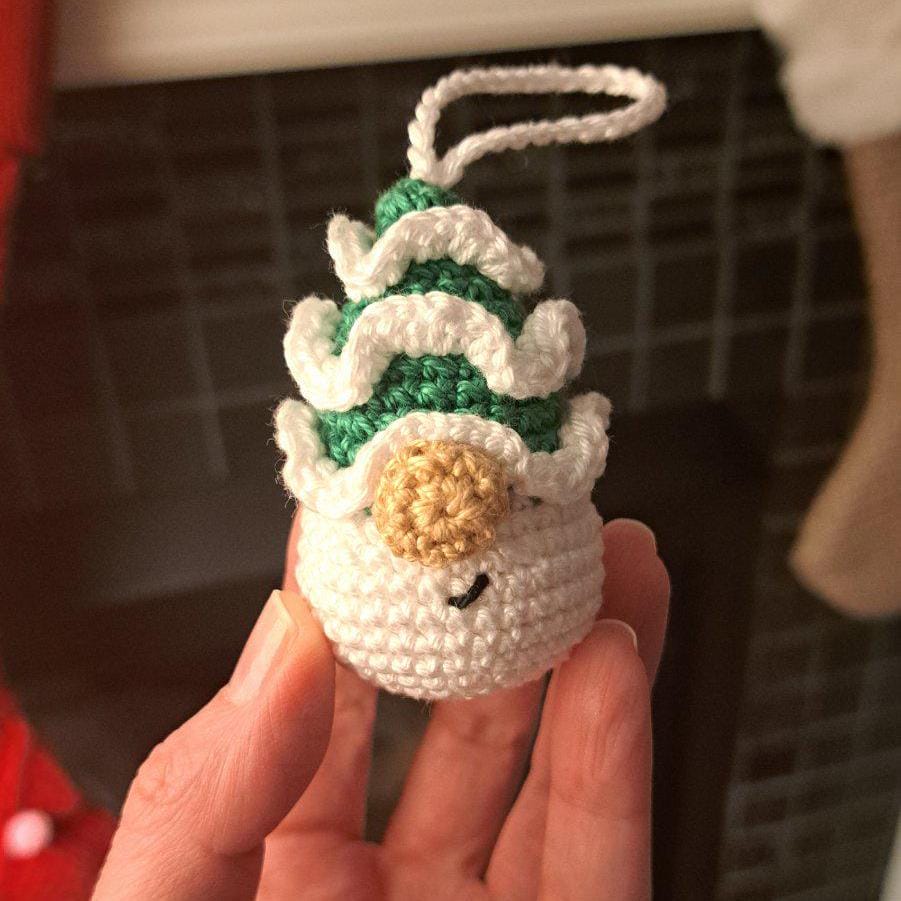 A small crocheted ornament shaped like a schristmsd tree gnome, held in a person's hand. The ornament is primarily white with green textured stripes, a beige round face, and a white hanging loop at the top.