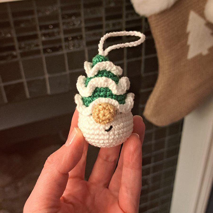 A hand holding a small crocheted Christmas ornament shaped like a snowman with a green and white hat, a beige nose, and a white hanging loop, against a dark tiled background with a stocking partially visible.