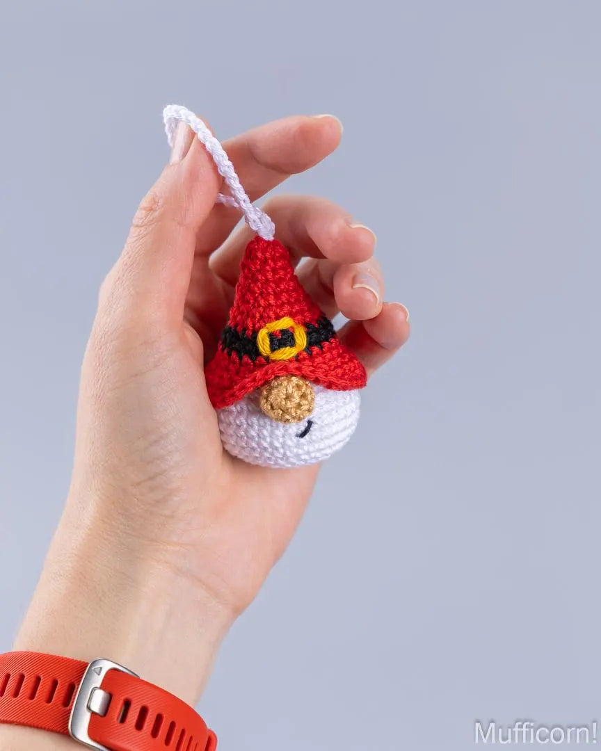 A hand holding a small crocheted gnome ornament with a red hat, black belt, yellow buckle, white face, and orange carrot nose against a plain light gray background.