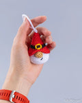 A hand holding a small crocheted gnome ornament with a red hat, black belt, yellow buckle, white face, and orange carrot nose against a plain light gray background.