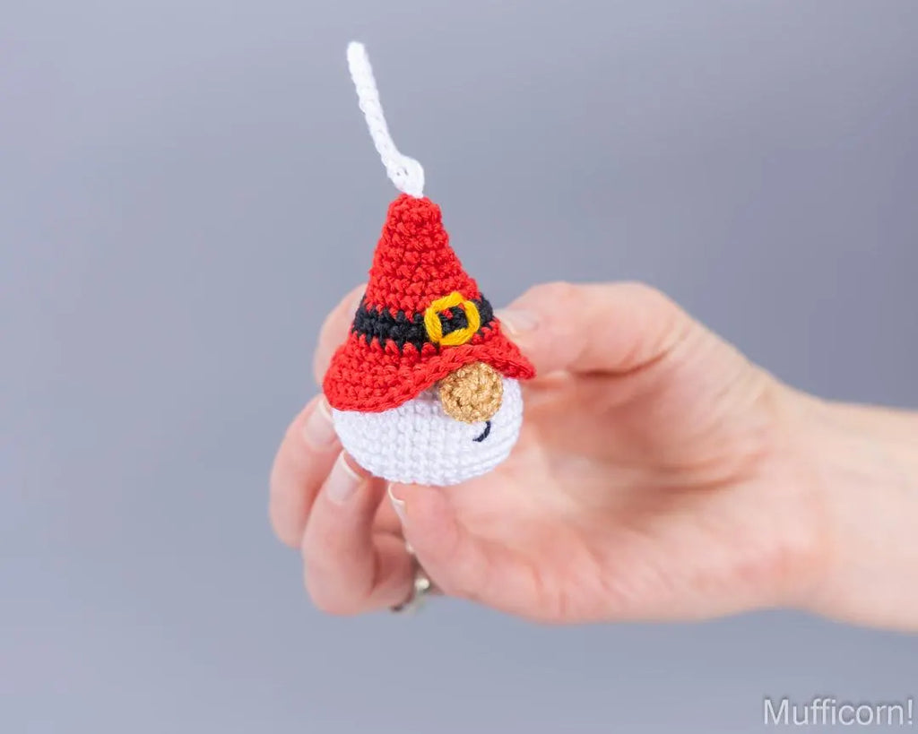 A hand holding a small crocheted ornament shaped like Santa, featuring a white brim, black band, yellow buckle detail, and a white hanging loop, against a plain gray background.