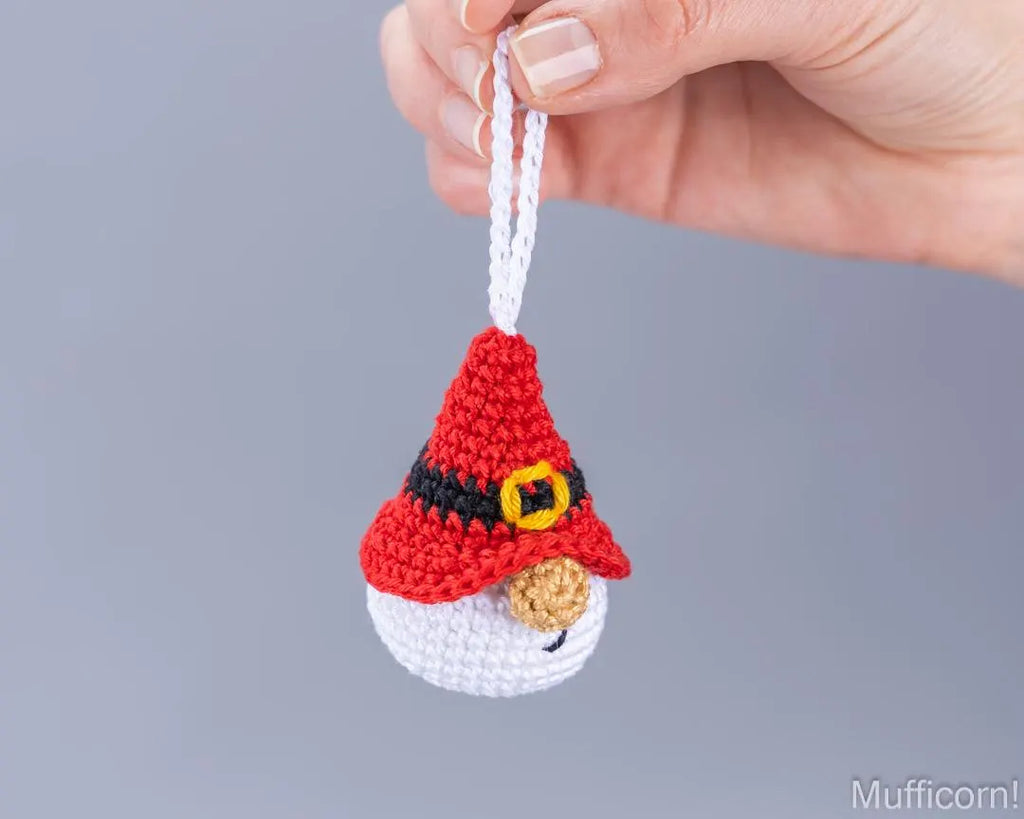 A hand holds a small crocheted muffin-shaped ornament with a red hat, black belt, yellow buckle, white beard, and white hanging loop against a plain gray background.