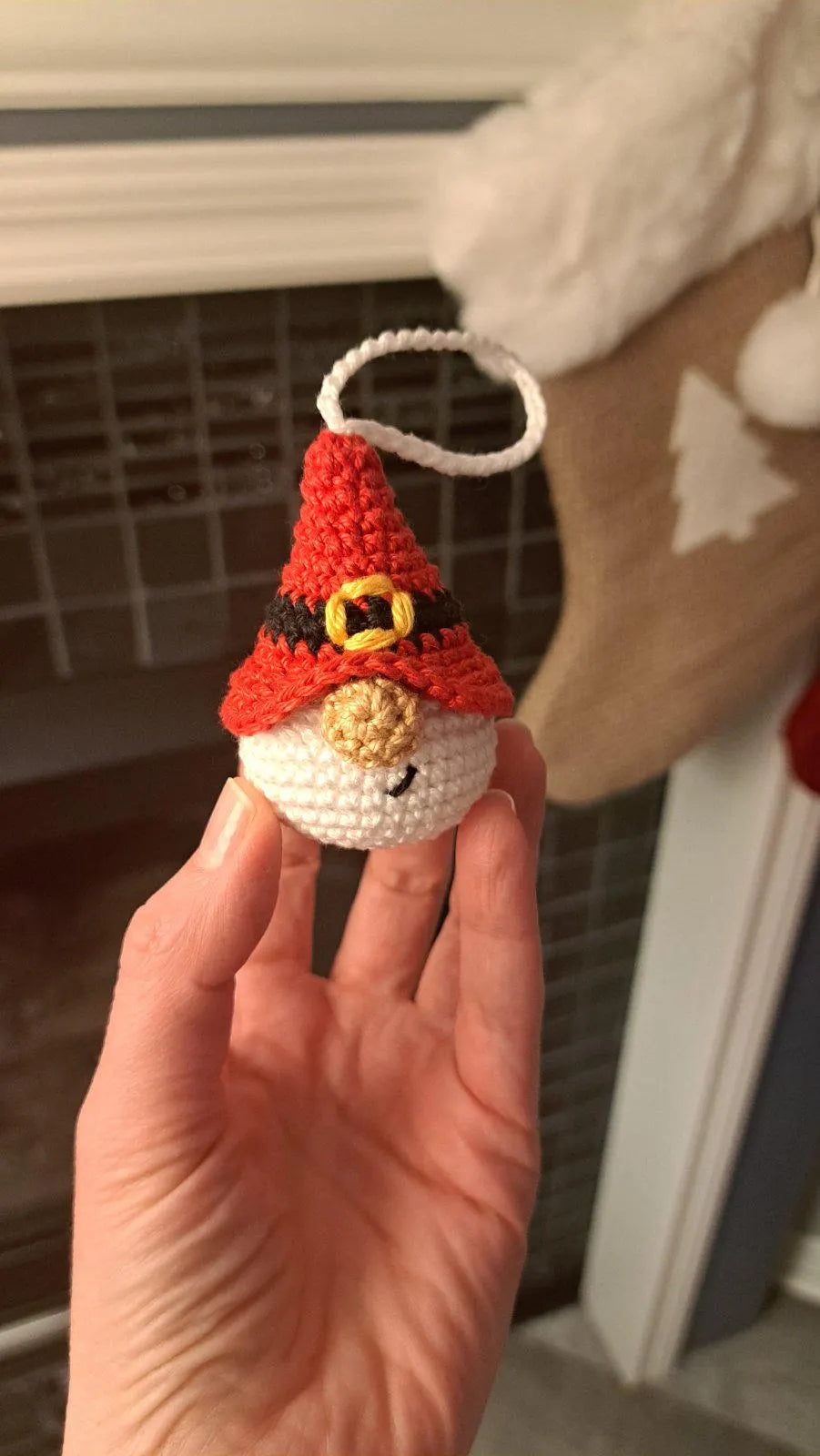 A hand holding a small crocheted Christmas gnome ornament with a red hat, white face and body, and a gold nose, shown in a home setting.