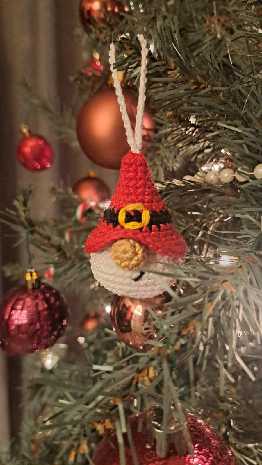 A handmade crocheted Christmas ornament shaped like a snowman wearing a red pointed hat with a black and gold band, hanging on a decorated Christmas tree with red and copper ornaments.