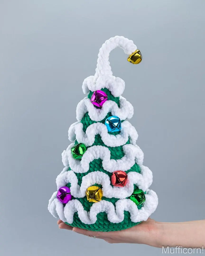 A crocheted Christmas tree ornament held in a hand against a plain gray background. The ornament is green and white with shiny multicolor gem-like ornaments and a gold bell at the top.