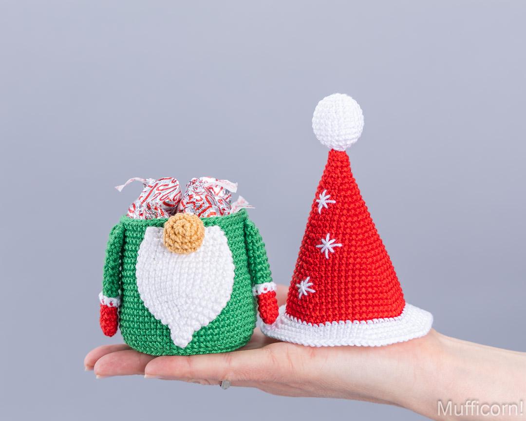 A crocheted Christmas gnome gift bag in green and white holding candy, displayed on a person's hand next to a crocheted red Santa hat with white trim and pom-pom.