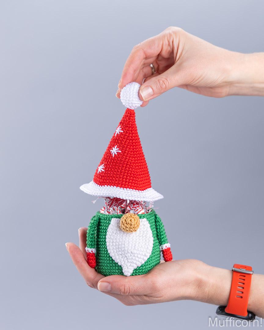 A person's hands holding a small crocheted Christmas gnome figurine, with one hand adjusting its red Santa hat. The gnome has a green body, a white beard, and a gold nose, with candy visible inside its head.