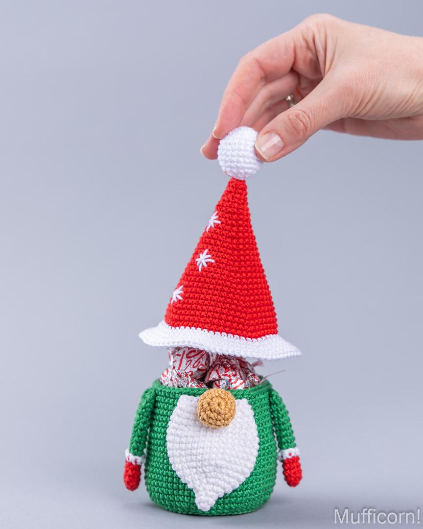 A crocheted Christmas gnome candy holder filled with wrapped chocolates, with a hand holding the pointed red Santa hat above it, against a plain light gray background.