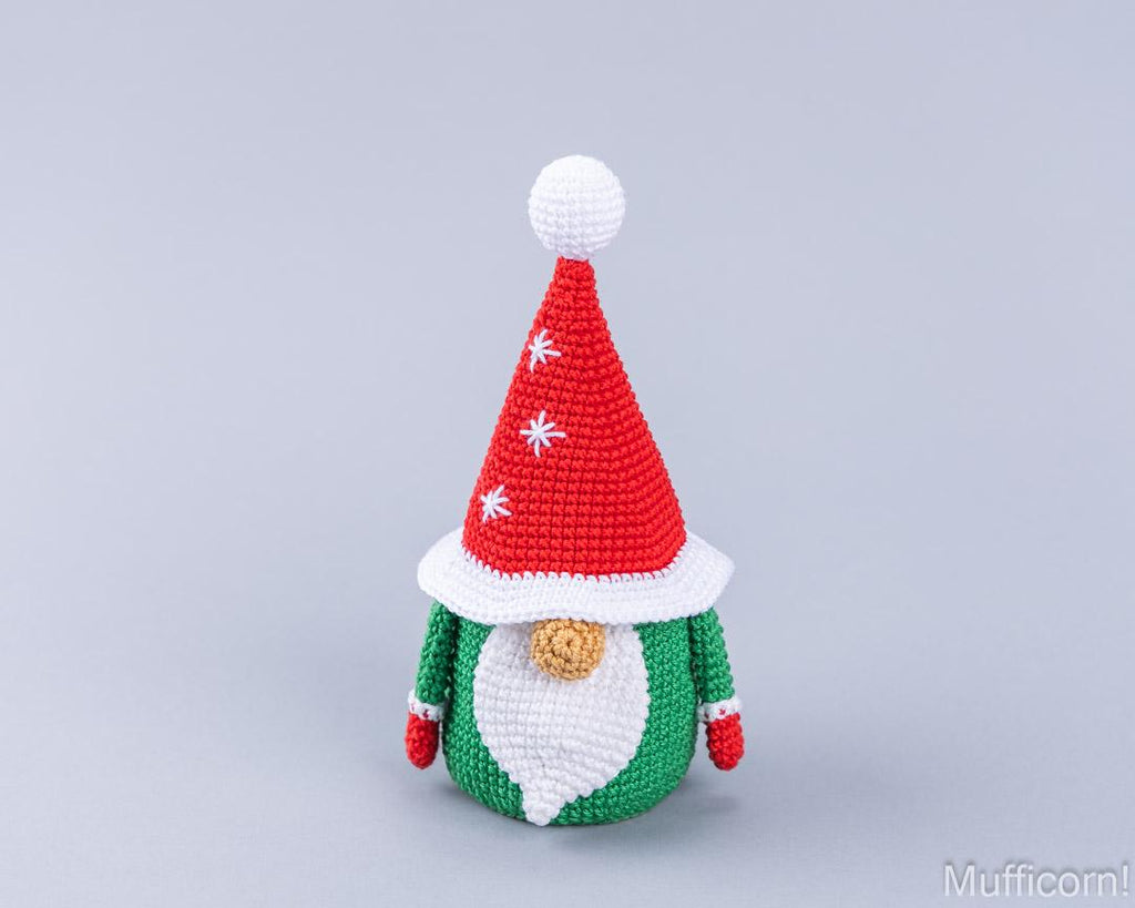 A crocheted Christmas gnome doll with a red pointed hat, white pom-pom and trim, green body, white beard, and small gold nose, centered on a light gray background.