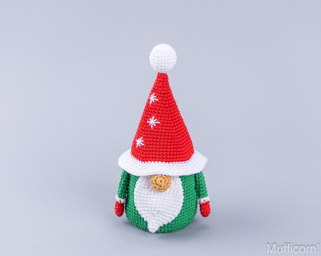 A crocheted Christmas gnome doll with a red pointed hat, white pom-pom and trim, green body, white beard, and small gold nose, centered on a light gray background.