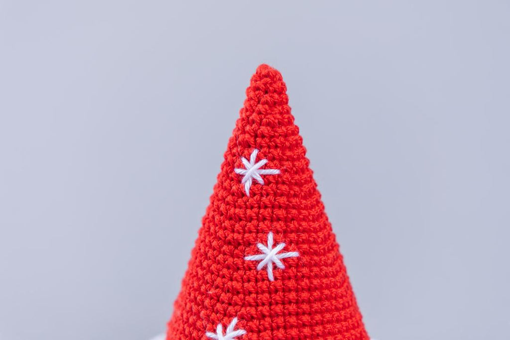 A close-up of a red crocheted cone-shaped item, likely a decorative cone or hat, decorated with three white embroidered snowflake patterns, set against a solid light gray background.