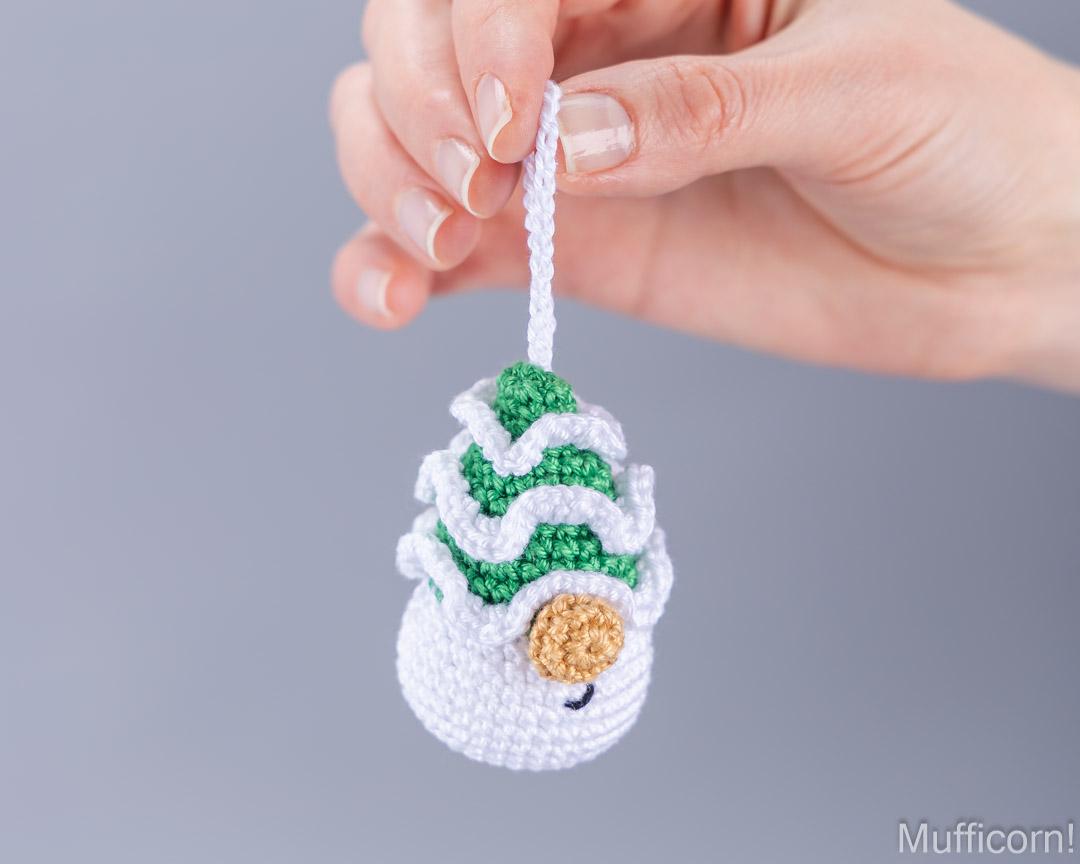 A hand holds a small crocheted muffin ornament against a plain gray background. The ornament is white with green frosting swirls, a tan crumb topping detail, and a white hanging loop.