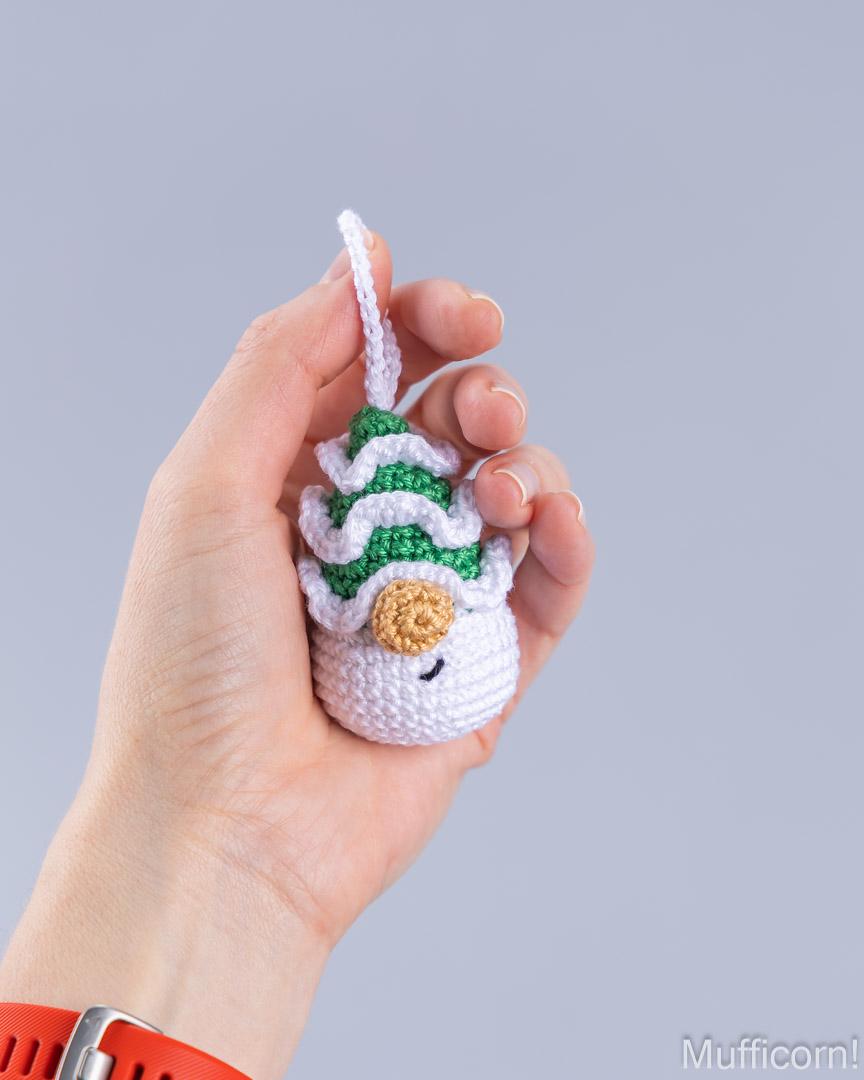 A hand holding a small crocheted snowman ornament with a green and white striped hat and a tan carrot nose against a solid gray background.