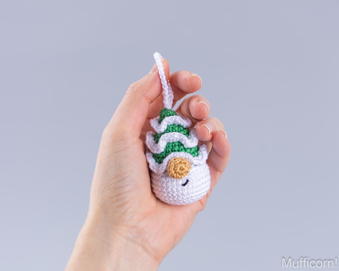 A hand holding a small christmas tree gnome ornament with a green and white striped hat and a tan button, against a plain light gray background.