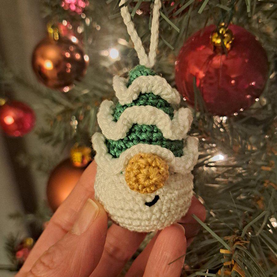 A handmade crocheted Christmas ornament shaped like a christmas tree wearing a green and white ruffled hat, held in a hand in front of a decorated Christmas tree with red and gold ornaments.