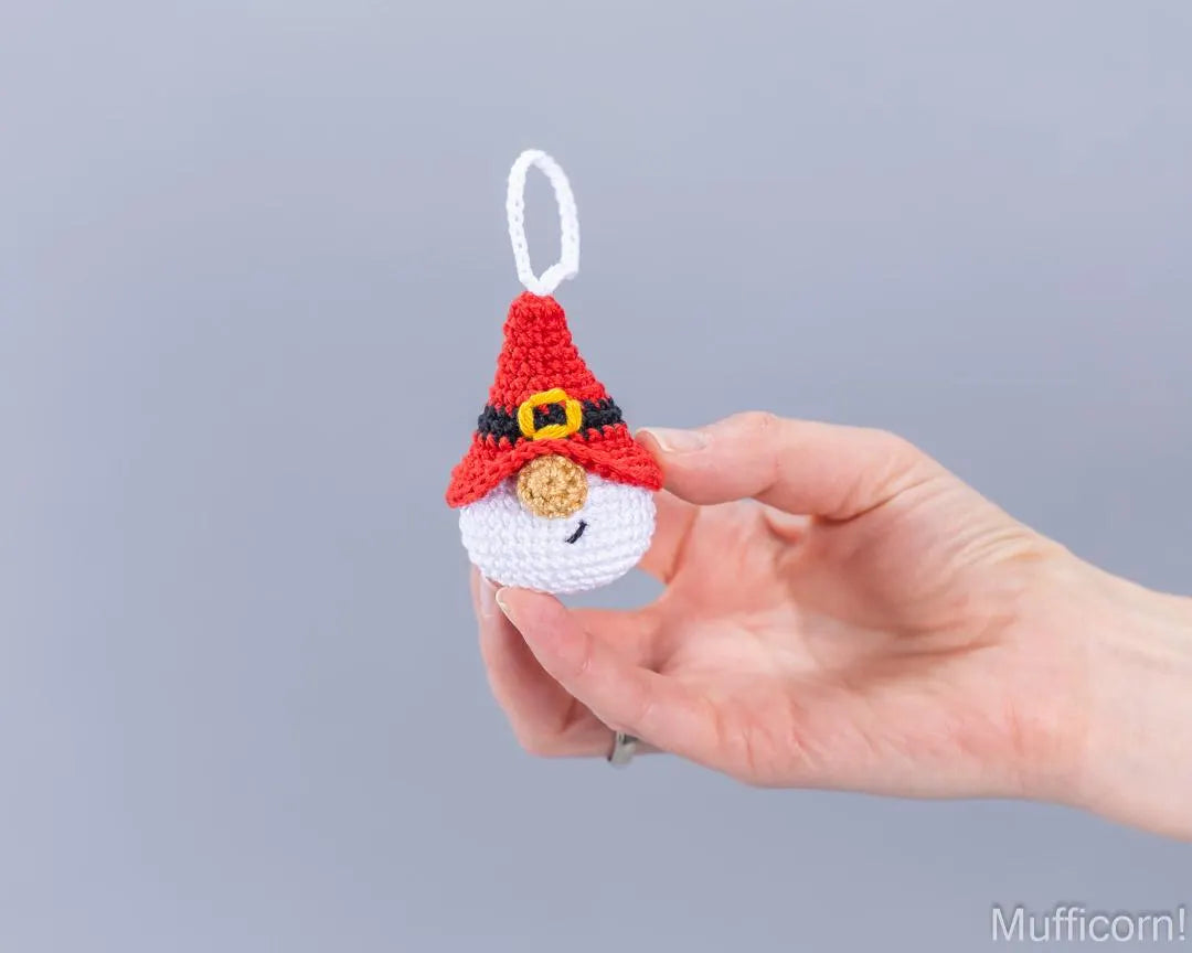 A hand holding a small crocheted gnomehorn ornament against a plain gray background. The ornament features a red hat with a black band and gold buckle, a white beard, and a white hanging loop.