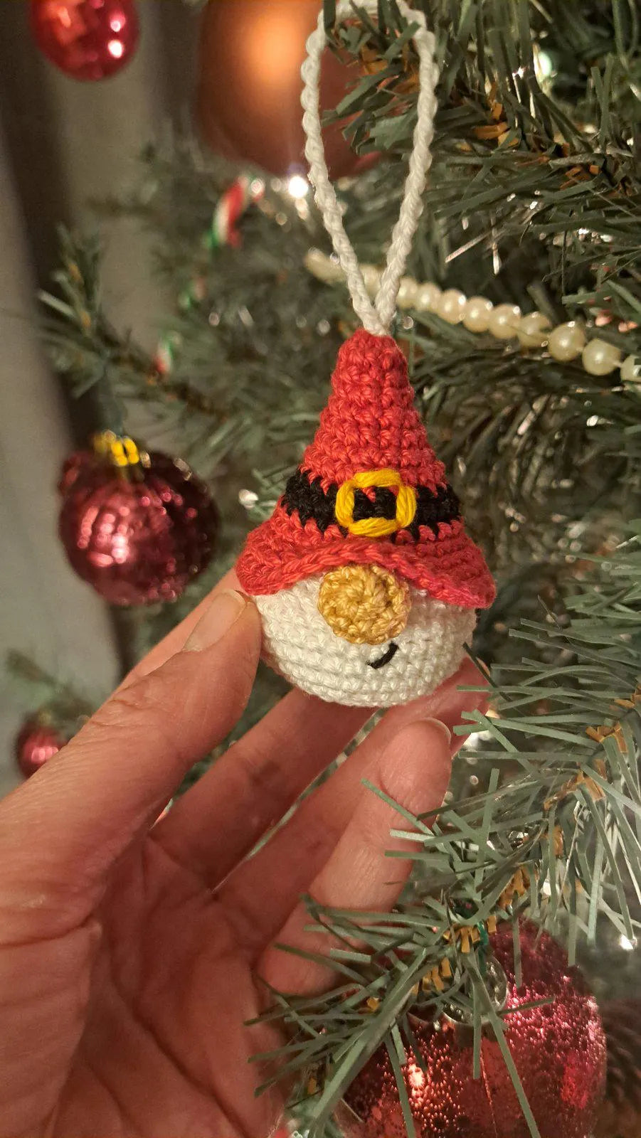 A hand holds a small crocheted Christmas ornament shaped like a gnome or snowman character wearing a red hat with a yellow and black buckle detail, hanging against a decorated Christmas tree with red ornaments.