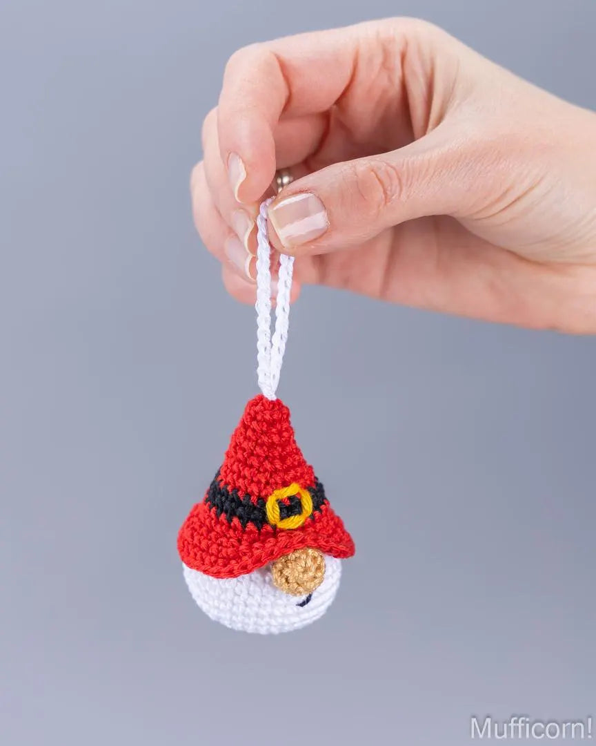 A hand holding a small crocheted Santa hat ornament against a plain gray background. The ornament features red, white, black, yellow, and brown details, with a white hanging loop.