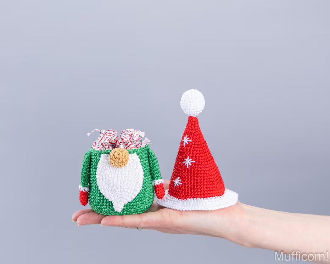 A hand holds two crocheted Christmas-themed accessories: a green gift bag with a white beard and red mittens, and a red Santa hat with white trim and pom-pom.