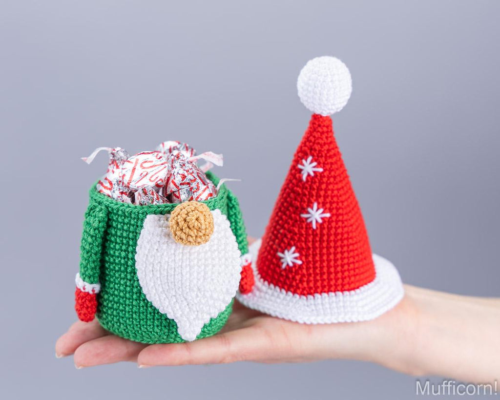 A crocheted Christmas elf gift basket and Santa hat displayed on a person's hand against a solid gray background.