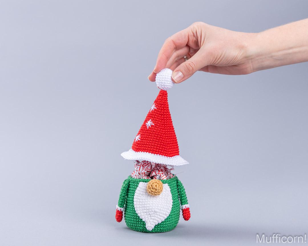 A hand is holding the top of a crocheted Christmas gnome doll wearing a red Santa hat with white trim and a green body with a white beard, against a plain light gray background.