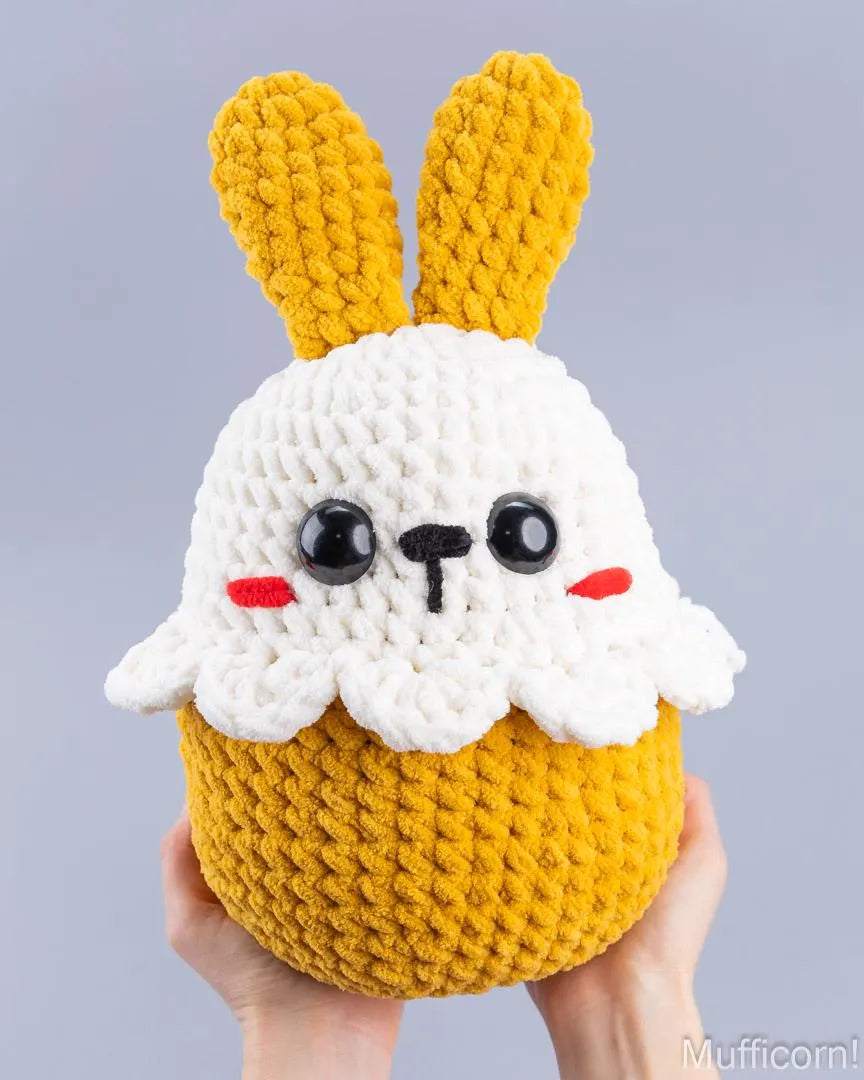 A crocheted plush bunny cupcake held in two hands against a solid light gray background. The plush toy features a white yarn bunny head with black eyes, red cheeks, and a yellow yarn body and ears.