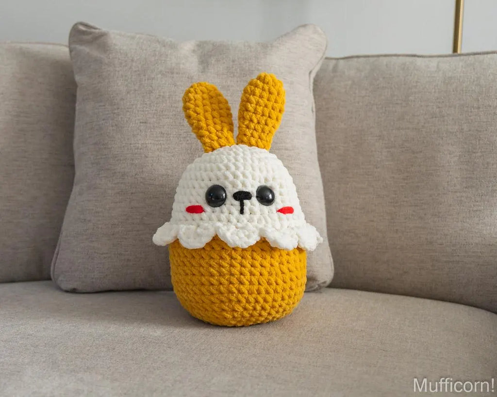A crocheted plush bunny pillow with long yellow ears and a white face sits on a beige sofa in front of a matching pillow. The bunny has black embroidered eyes and nose with small red cheeks.