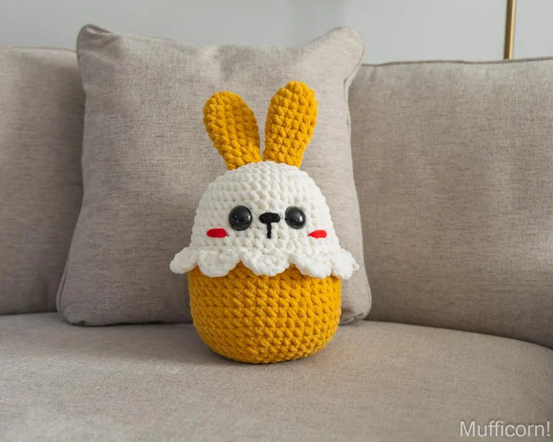 A crocheted plush bunny pillow with long yellow ears and a white face sits on a beige sofa in front of a matching pillow. The bunny has black embroidered eyes and nose with small red cheeks.