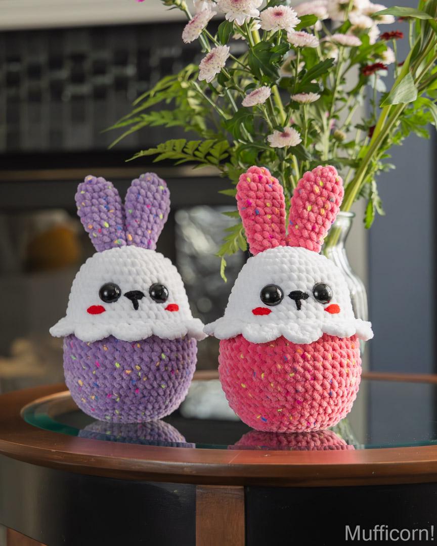 Two crocheted bunny sitting on a glass table, one in purple and one in pink, with a vase of flowers behind them.