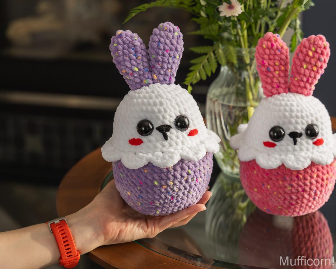 Two handmade crocheted bunny, one purple and one pink, displayed on a glass table with a hand holding the purple one. A glass vase with flowers is in the background.