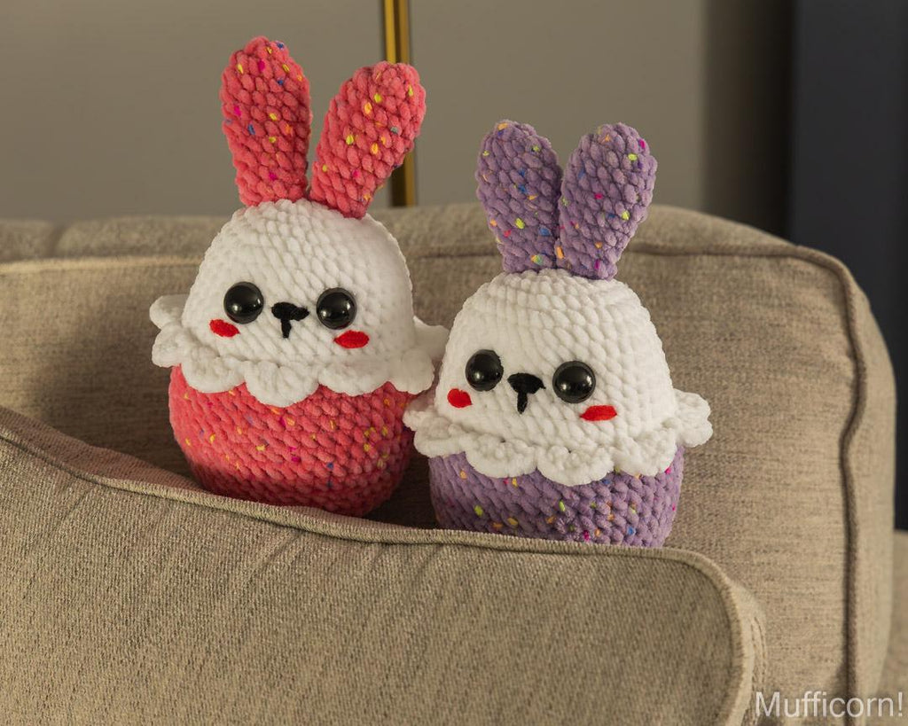 Two handmade crocheted bunny cupcake toys sitting on a beige fabric armchair. The toy on the left is pink and white, and the toy on the right is purple and white, both with multicolored yarn details on the ears and cupcakes.
