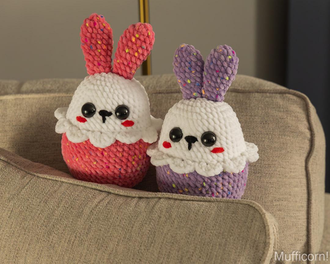 Two handmade crocheted bunny cupcake toys sitting on a beige fabric armchair. The toy on the left is pink and white, and the toy on the right is purple and white, both with multicolored yarn details on the ears and cupcakes.