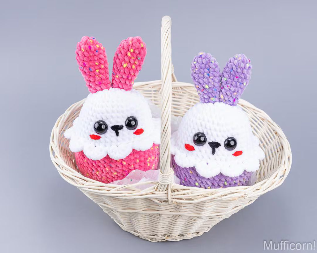 Two cute crocheted bunny plush toys sitting in a white wicker basket. One bunny is pink and white, and the other is purple and white, both with black eyes and red cheeks, set against a solid gray background.