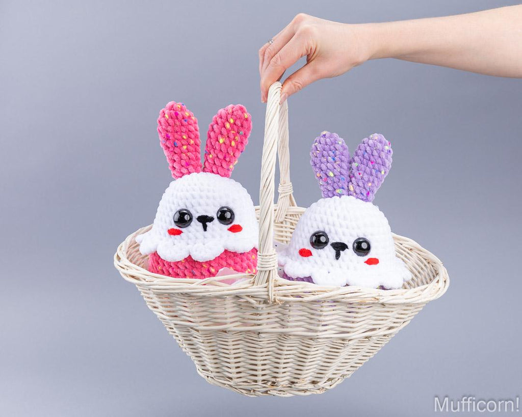 Two crocheted bunny figures sitting in a woven basket, one with pink ears and one with purple ears, held by a person's hand against a plain gray background.