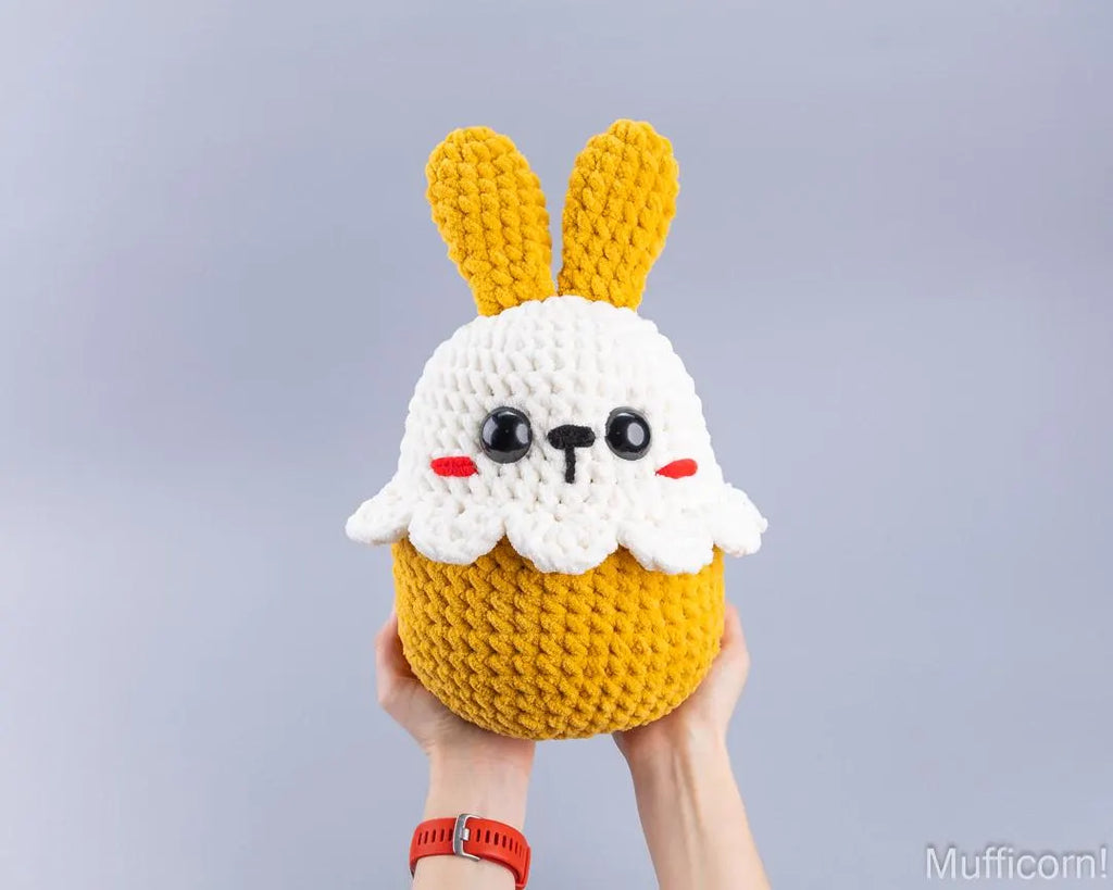 A handmade crocheted plush toy shaped like a cute bunny head on a yellow cupcake, held up by two hands against a plain light gray background.