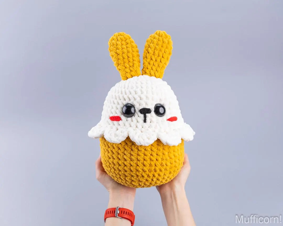 A handmade crocheted plush toy shaped like a cute bunny head on a yellow cupcake, held up by two hands against a plain light gray background.