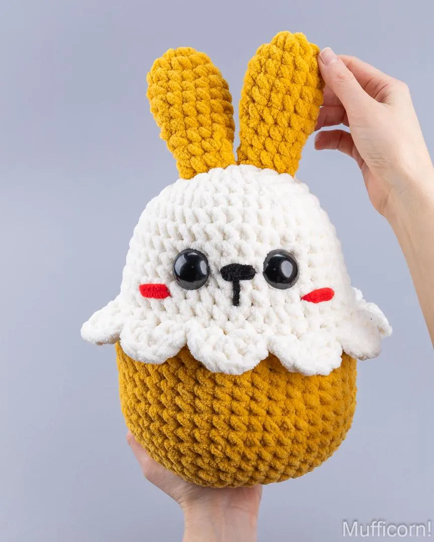 A handmade crocheted plush bunny cupcake toy being held by two hands against a solid light gray background. The toy features a yellow-gold textured body and ears, a white textured frosting-like top, black embroidered eyes and nose, and small red embroidered cheeks.
