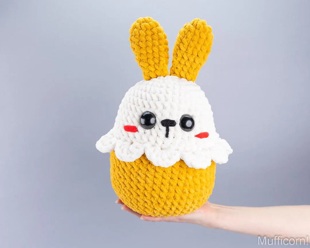 A hand holds a crocheted plush bunny cupcake toy against a plain light gray background. The toy has yellow yarn ears, a white yarn body, and a yellow yarn base, with embroidered black eyes, a black nose, and red cheeks.