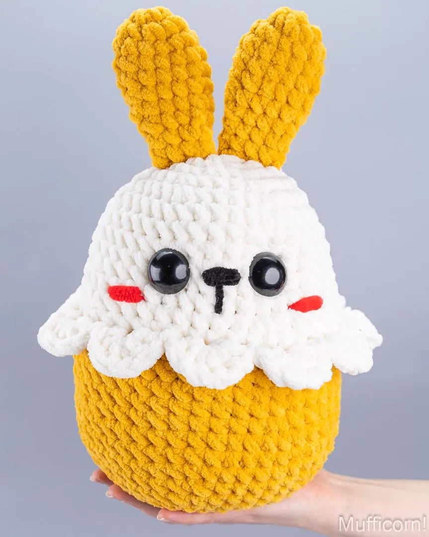 A handmade crocheted plush toy shaped like a cute bunny cupcake, held in a person's hand against a plain gray studio background. The plush has a white yarn body, yellow yarn ears and lower half, black embroidered eyes and nose, and small red embroidered cheeks.