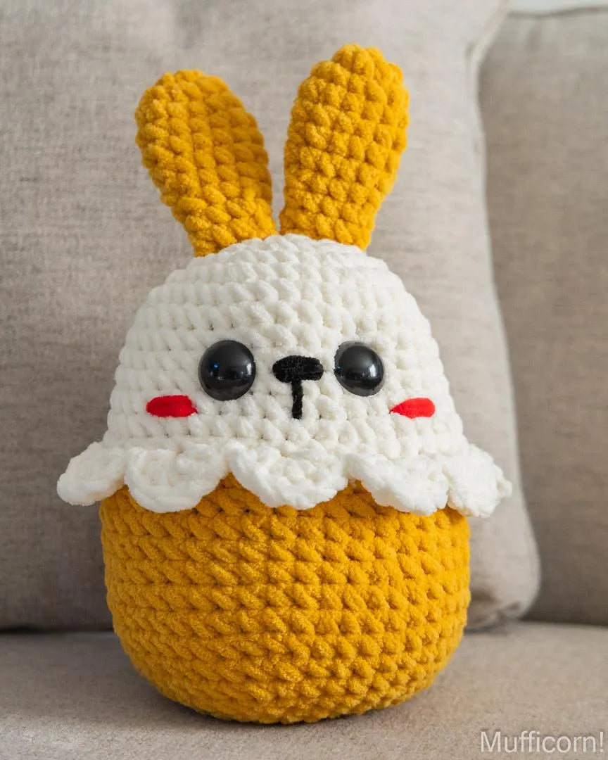 A crocheted plush bunny cupcake toy with a yellow body, white frosting-like top, and long yellow bunny ears, sitting on a beige fabric surface.