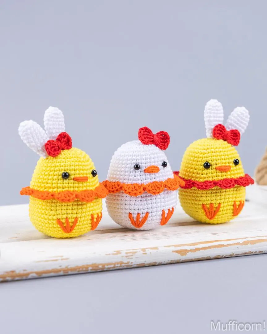 Three crocheted Easter duck figures are displayed on a distressed white wooden surface against a soft gray background. The left and right figures are yellow with white bunny ears, red bows, orange ruffles, and orange feet.
