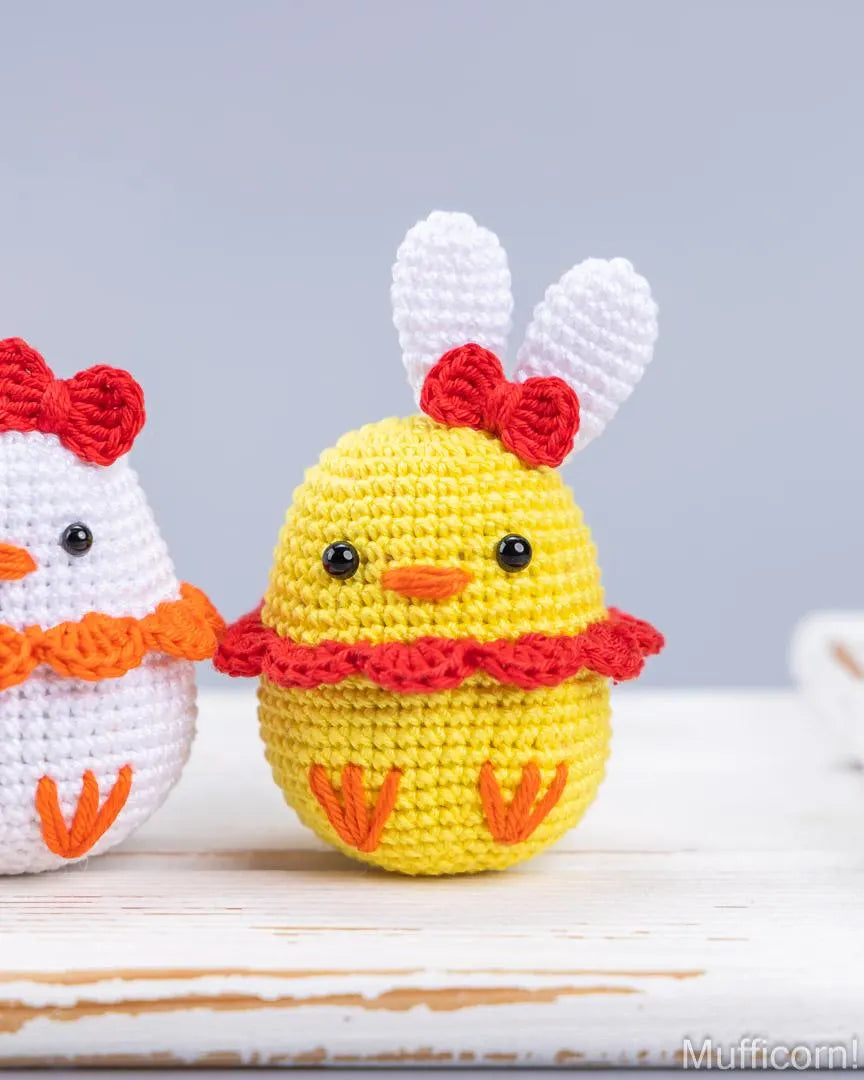 A crocheted Easter egg duck toy in bright yellow with white bunny ears, a red bow, and a red ruffled collar, standing on a white wooden surface. A second crocheted egg character is partially visible on the left.