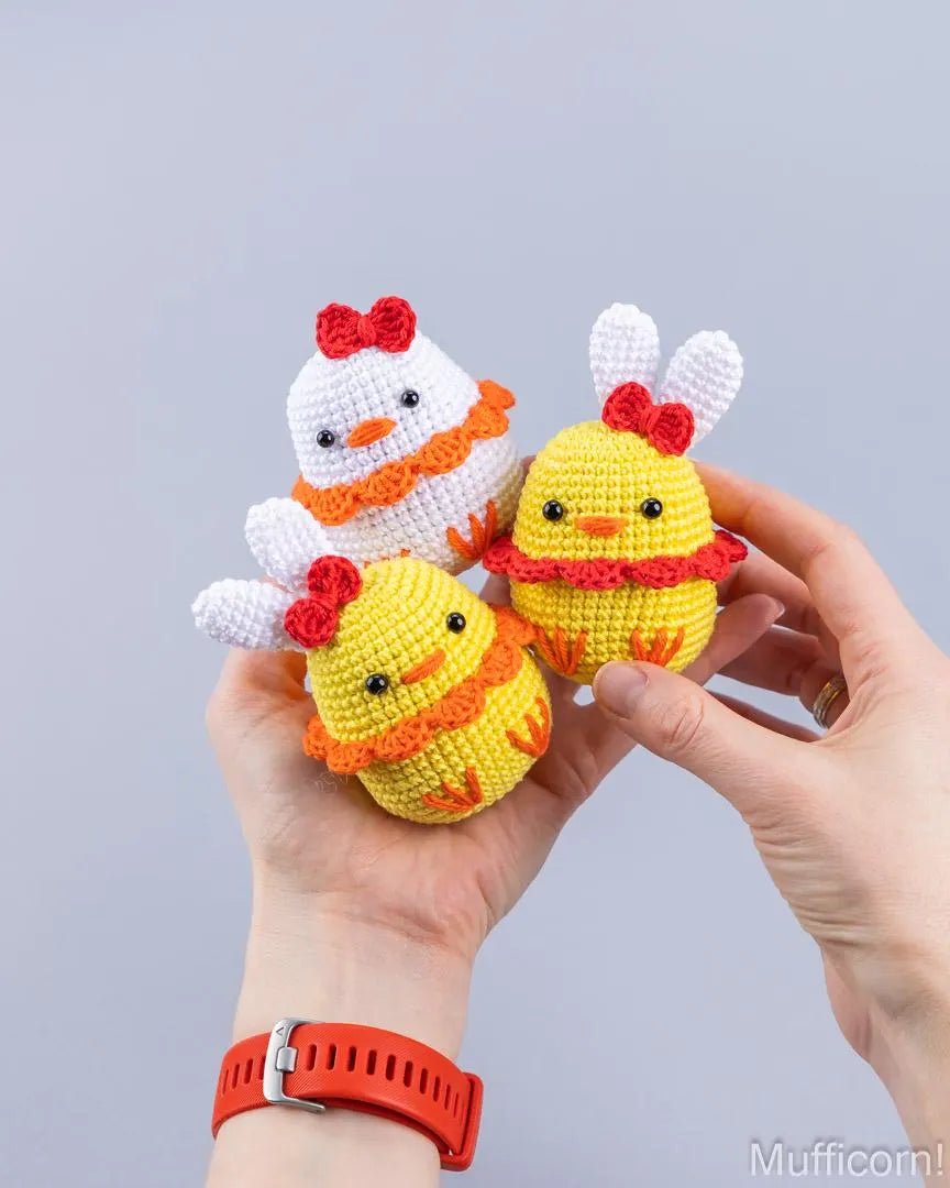 Three crocheted amigurumi ducks in yellow, white, and yellow-orange are held in two hands against a light gray background. The chicks have embroidered eyes, beaks, and decorative bows and scarves.