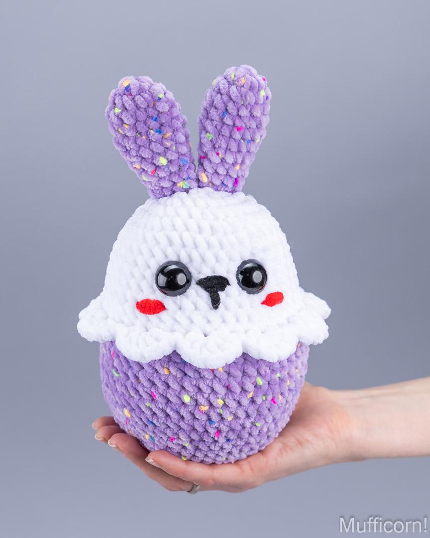 A handmade crocheted plush bunny cupcake toy held in a person's hand against a solid gray studio background. The toy features a white knitted top with black eyes, red cheeks, and a purple knitted bottom with bunny ears.